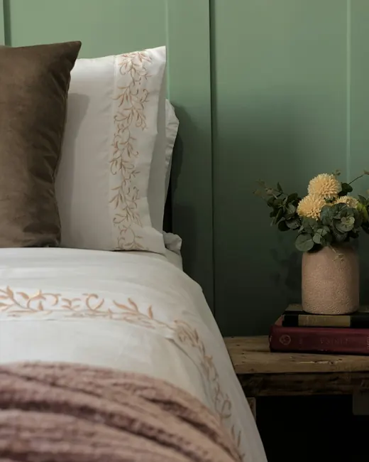 Lit au linge brodé contre un mur vert d’eau, bouquet de fleurs sèches sur la table de nuit
